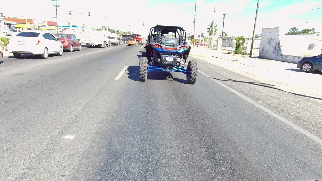 Blue UTV remains centered on the Los Cabos multi-lane road, with traffic ahead, a dark car near the right side, overexposed sidewalk areas, and no visible people.