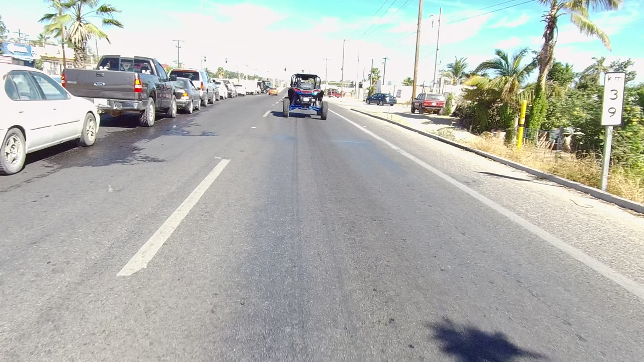 Opening frame of a Los Cabos multi-lane road with parked cars on the left, a small blue vehicle ahead, palm trees, sidewalk, clear sky, and no visible people.