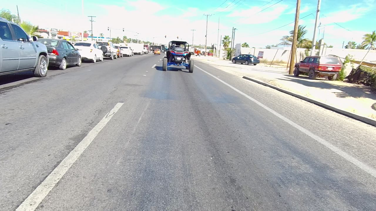 Mid-clip frame on a bright Los Cabos street, showing lane markings in the foreground, the small blue vehicle ahead, parked cars at left, a sidewalk and palms at right, and no visible people.