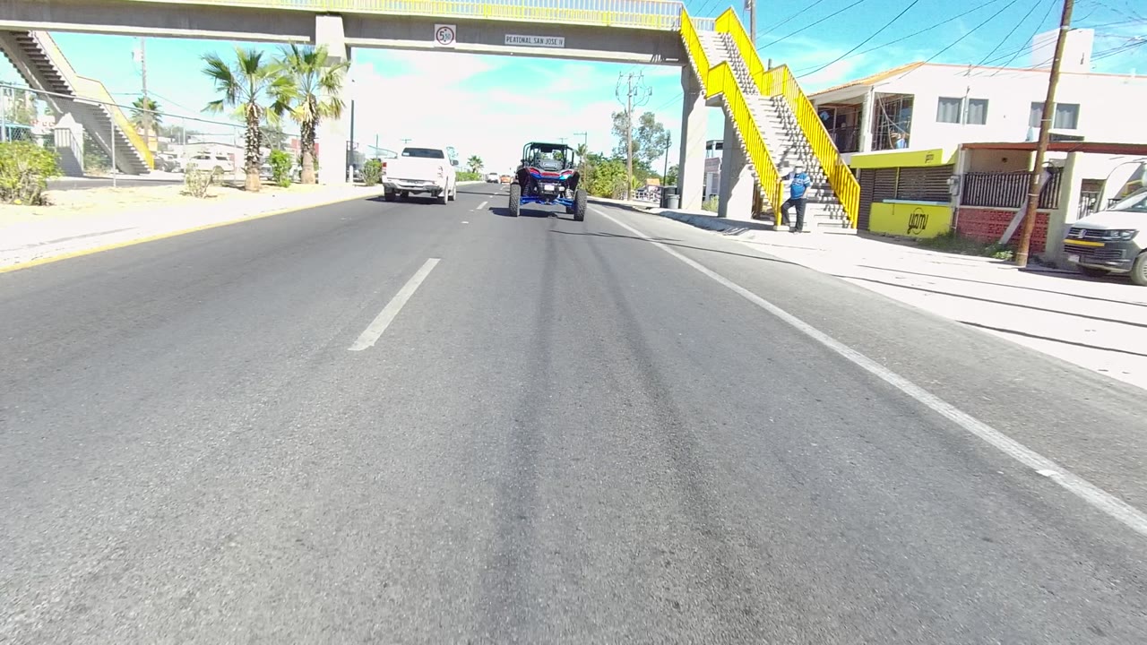 Forward road view in Los Cabos, Baja California Sur, approaching a yellow pedestrian overpass with a blue off-road vehicle ahead, roadside buildings, palm trees, and no visible people.