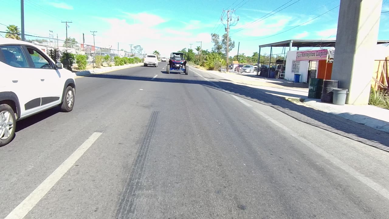 Street view in Los Cabos, Baja California Sur, with a white SUV close on the right near the yellow overpass stairs, traffic ahead, roadside buildings, and no visible people.