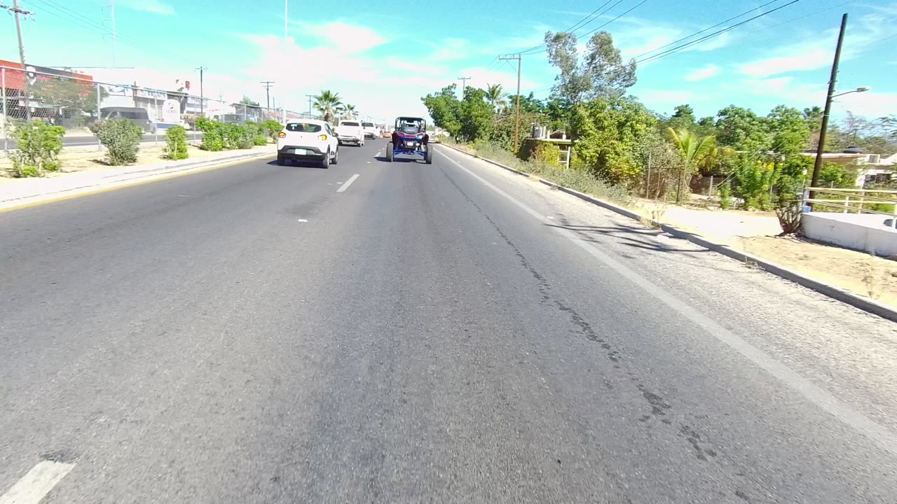 Opening frame of vehicles moving along a paved road in Los Cabos, Baja California Sur, with white lane markings, roadside bushes, utility poles, and no visible people.