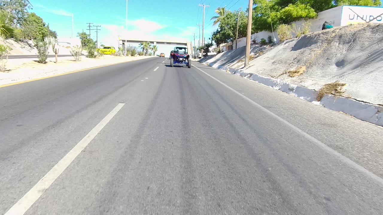 Forward view down a Los Cabos multi-lane road with a small off-road vehicle ahead, low buildings, palm trees, utility poles, bright sun, blue sky, and no visible people.