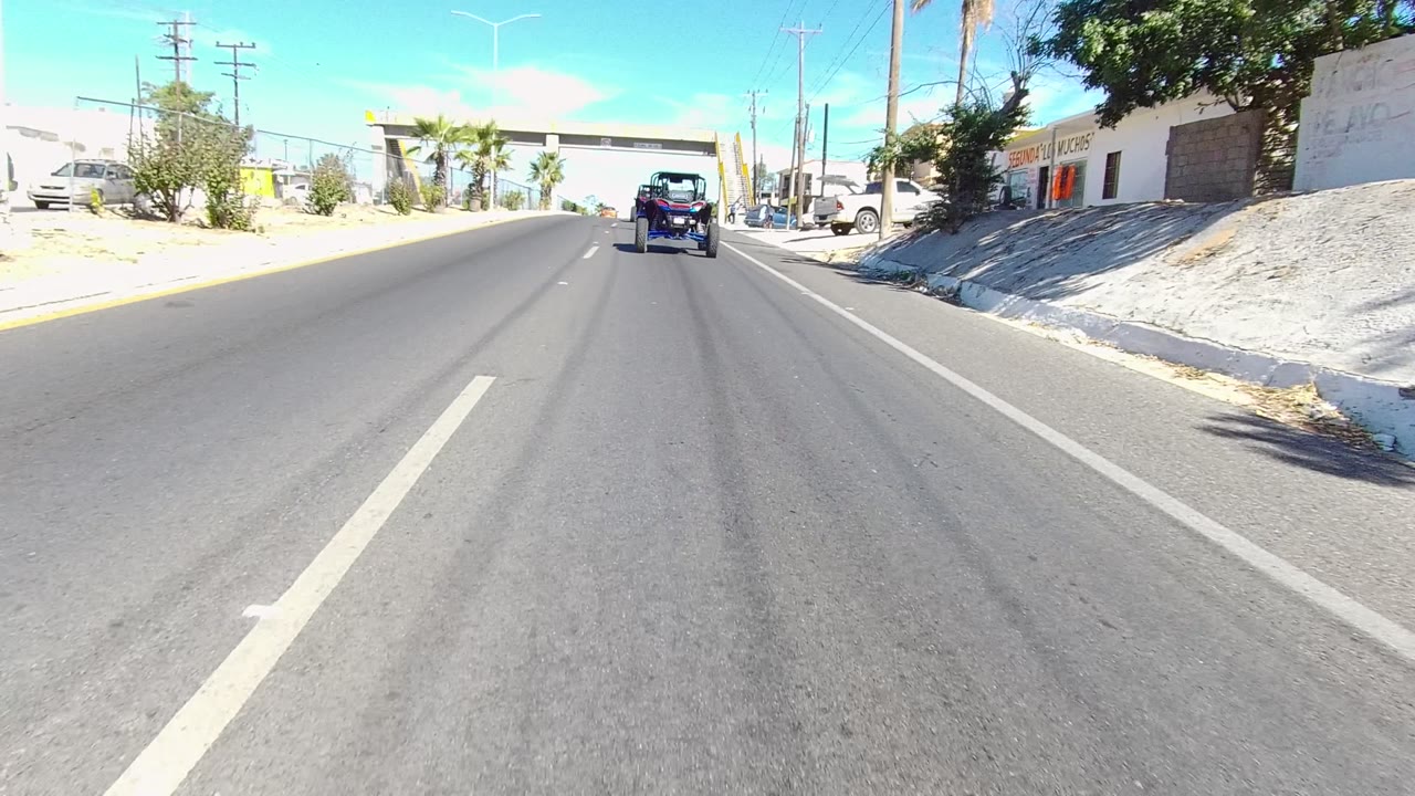 Forward Los Cabos street view with the small vehicle farther ahead, low commercial buildings, palm trees, utility poles, bright pavement, blue sky, and no visible people.