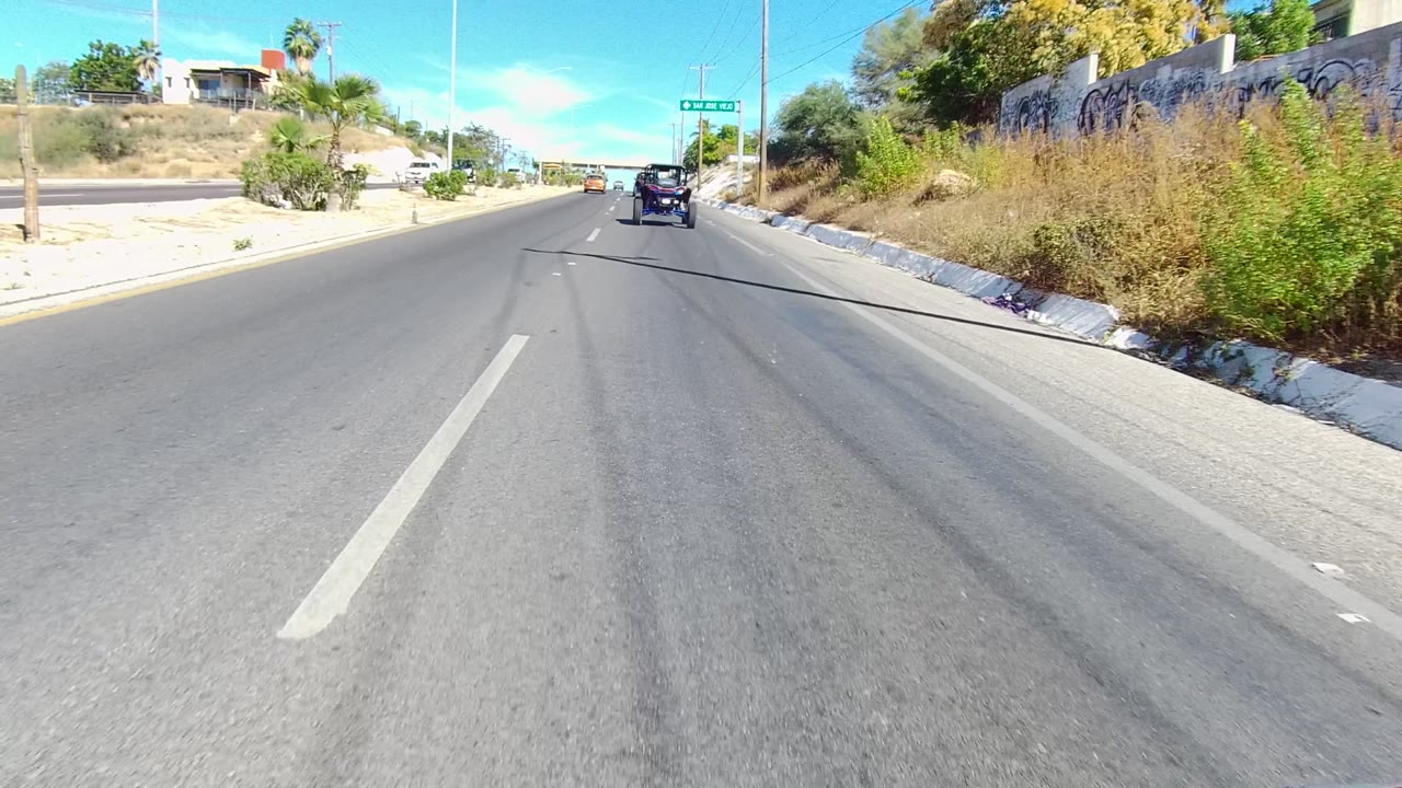 Forward road view in Los Cabos, Baja California Sur, Mexico, with a dark vehicle ahead, lane markings, dry roadside vegetation, palms, low buildings, and no visible people.