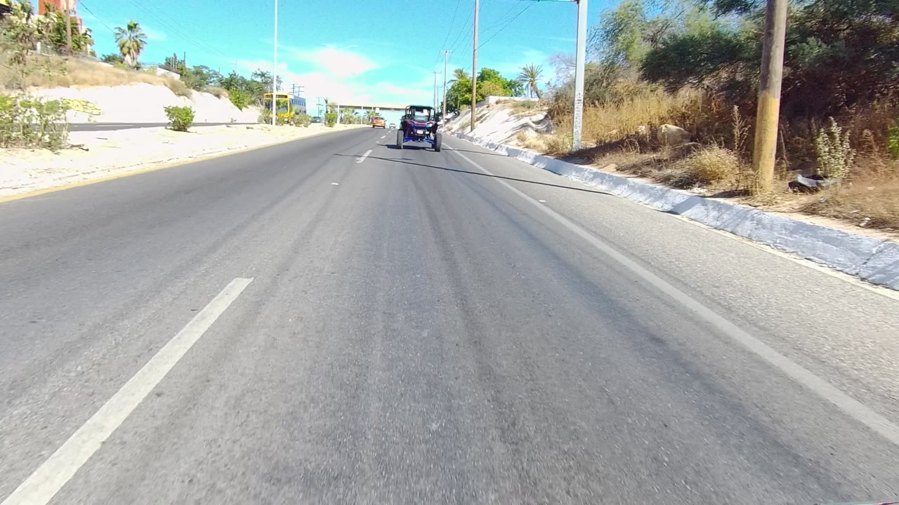 Forward-facing Los Cabos road view with a dark vehicle ahead, a side street on the right, dry brush, pale curbs, utility poles, palms, and no visible people.