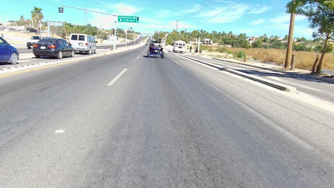 Forward road view in Los Cabos, Baja California Sur, with cars, a white van, a blue carriage-like vehicle ahead, street signs, palms, and no clearly visible people.