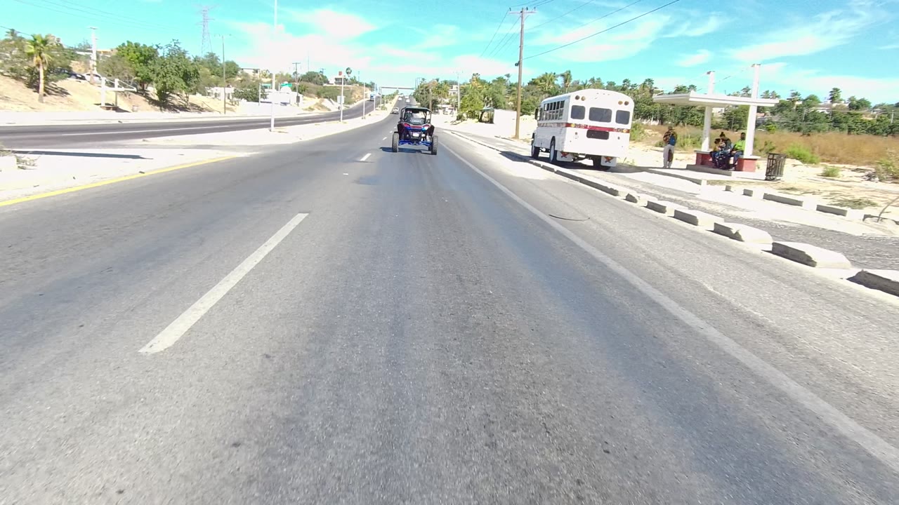 A Los Cabos road frame shows a white bus on the right, a bus shelter, green street signs, the blue carriage-like vehicle ahead, desert landscaping, and no clearly visible people.