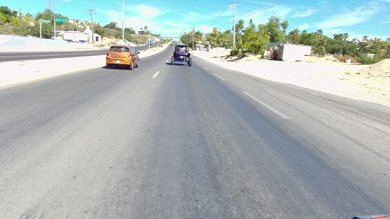 Forward road view in Los Cabos, Baja California Sur, with an orange car and dark off-road vehicle ahead, sandy shoulders, roadside greenery, low buildings, utility poles, and no visible people.