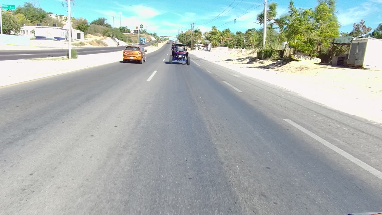 Two vehicles ahead on a paved road bordered by sand, trees, and buildings.