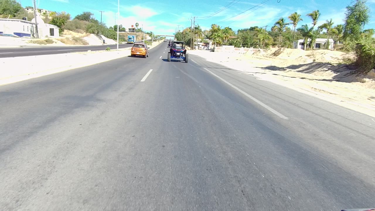 Forward-facing Los Cabos road view with the orange car and dark off-road vehicle ahead, sandy graded edges, roadside palms and shrubs, utility lines, low walls, and no visible people.