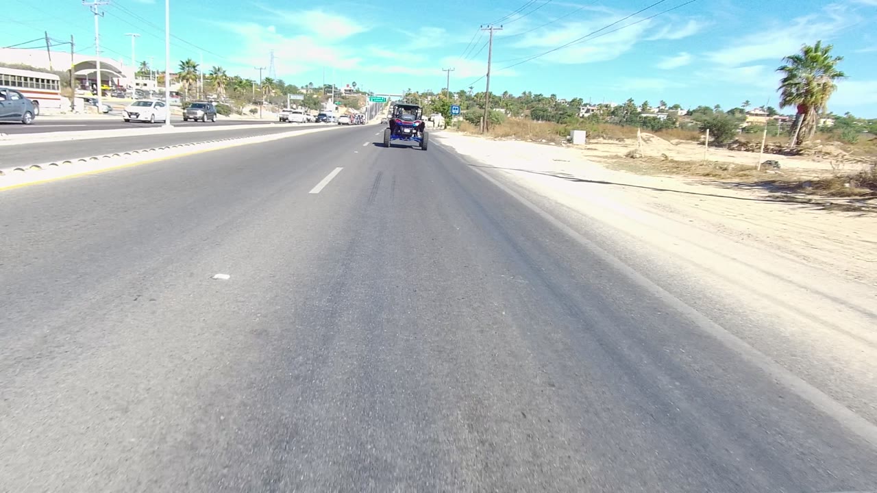Forward road view in Los Cabos, Baja California Sur, Mexico, with cars ahead, a dirt shoulder, utility poles, palm trees, and no visible people.