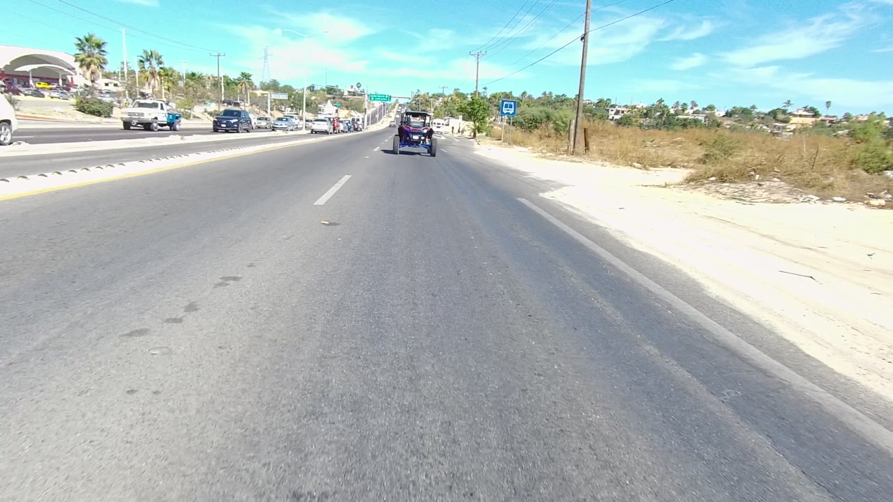 The road view in Los Cabos includes cars ahead, a blue bus stop sign on the right shoulder, dry brush, power lines, and no visible people.