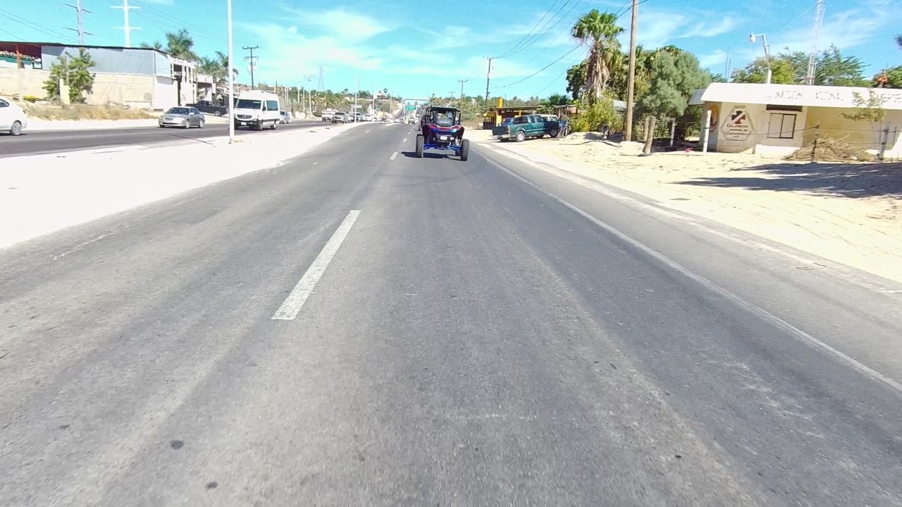 Los Cabos road-level view of a paved lane with a small cart ahead, cars on both sides, low roadside buildings, utility poles, sandy shoulders, and no visible people.