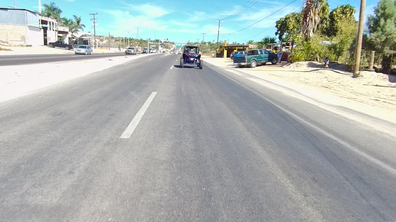 Forward road view with a cart ahead, parked vehicles, palms, and sandy roadside.