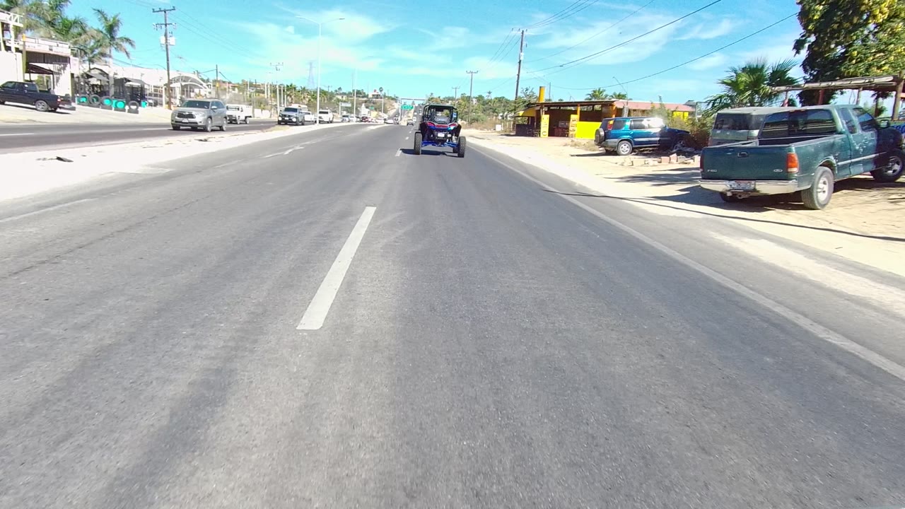 Los Cabos street frame with asphalt foreground, a small cart ahead in the lane, a green parked vehicle at the right shoulder, roadside trees, utility lines, and no visible people.