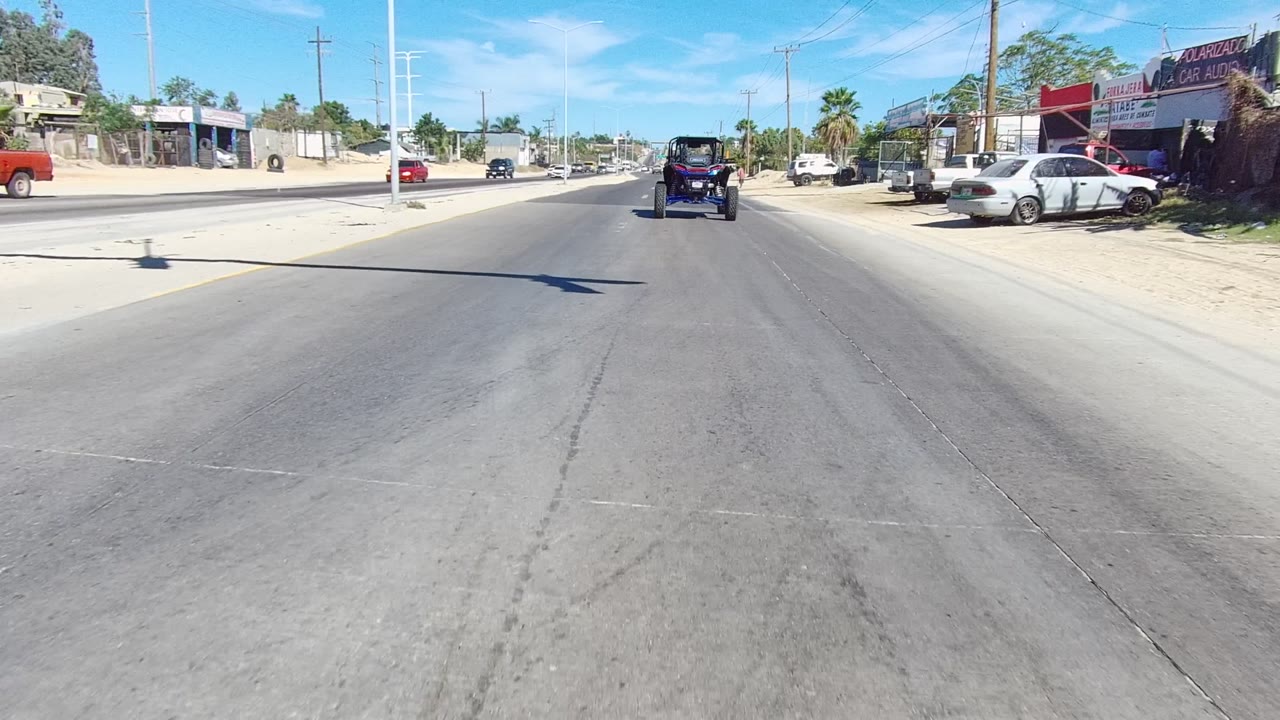 Street-level view in Los Cabos, Baja California Sur, showing a paved road, a dark off-road vehicle ahead, parked cars, business buildings, utility poles, sandy shoulders, and no visible people.