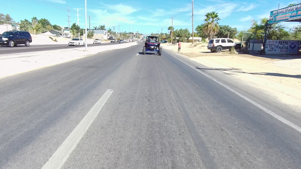Street view in Los Cabos with a wide paved road, a dark off-road vehicle ahead, a white truck parked near a roadside structure, sandy lots, utility poles, palm trees, and no visible people.
