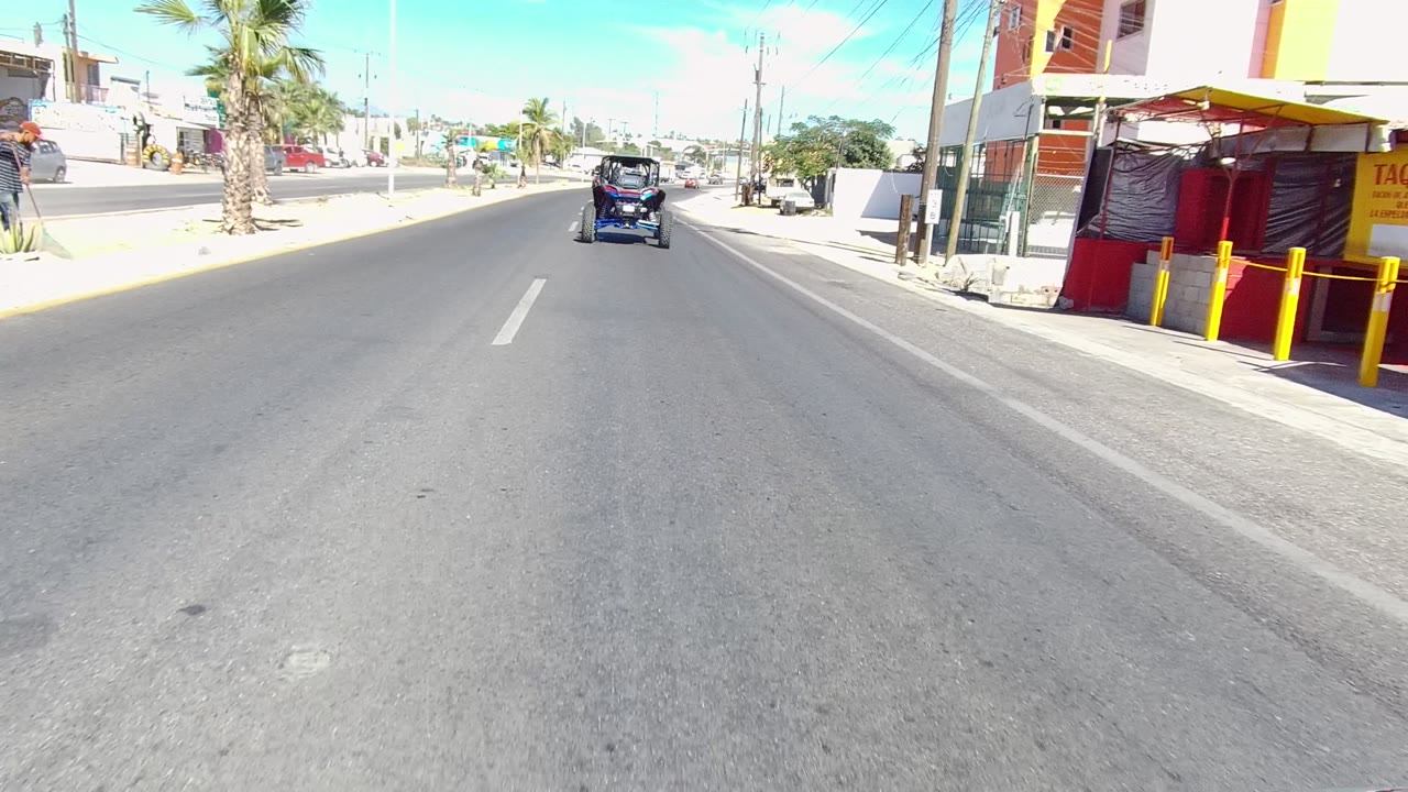 Start of a Los Cabos street-driving clip, showing a small vehicle ahead on a wide paved road with palms, storefronts, parked cars, utility poles, and no visible people.
