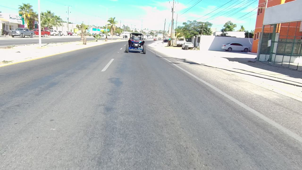 Small vehicle continues along the road beside a red roadside stand and palm trees.