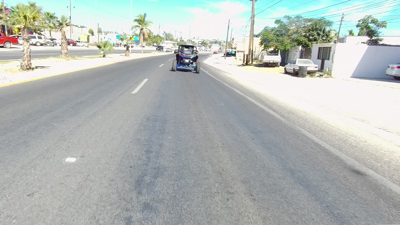 Mid-clip Los Cabos street frame with the small vehicle ahead, an orange building and fence to the right, parked cars, palm trees, utility lines, and no visible people.