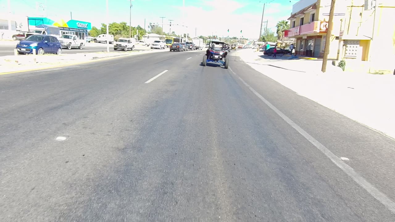 Road-level view in Los Cabos, Baja California Sur, Mexico, with a golf cart ahead, cars at both sides, storefronts, traffic lights, bright sun, and no clearly visible people.