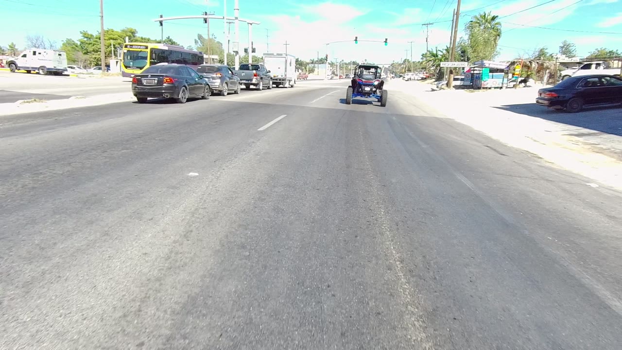 The Los Cabos roadway view shows the golf cart ahead, a dark car at left, a red storefront on the right, traffic signals overhead, palm trees, and a small distant person near the sidewalk.