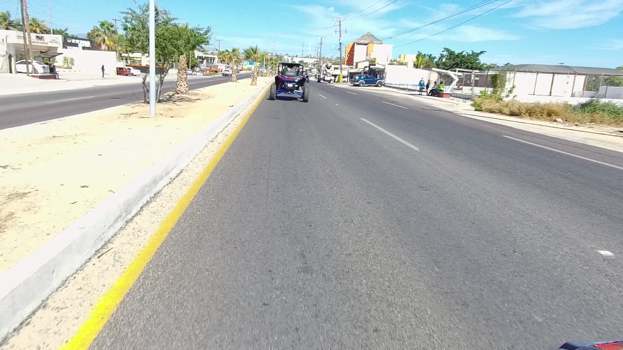 Forward road view in Los Cabos, Baja California Sur, with a dark off-road vehicle ahead, a yellow curb line, low roadside buildings, palms, and bright blue sky.