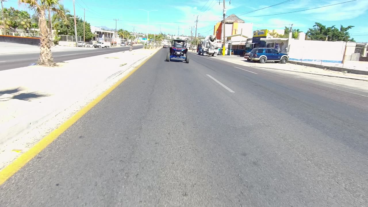 In Los Cabos, Baja California Sur, the camera view passes a roadside business area with parked vehicles, yellow posts, a cement mixer truck on the right, palms, and utility lines.