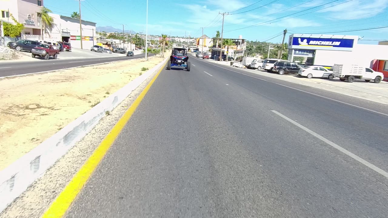 A golf cart moves ahead on a paved road in Los Cabos, Baja California Sur, with a sandy median curb, parked cars, commercial buildings, clear sky, and no visible people.