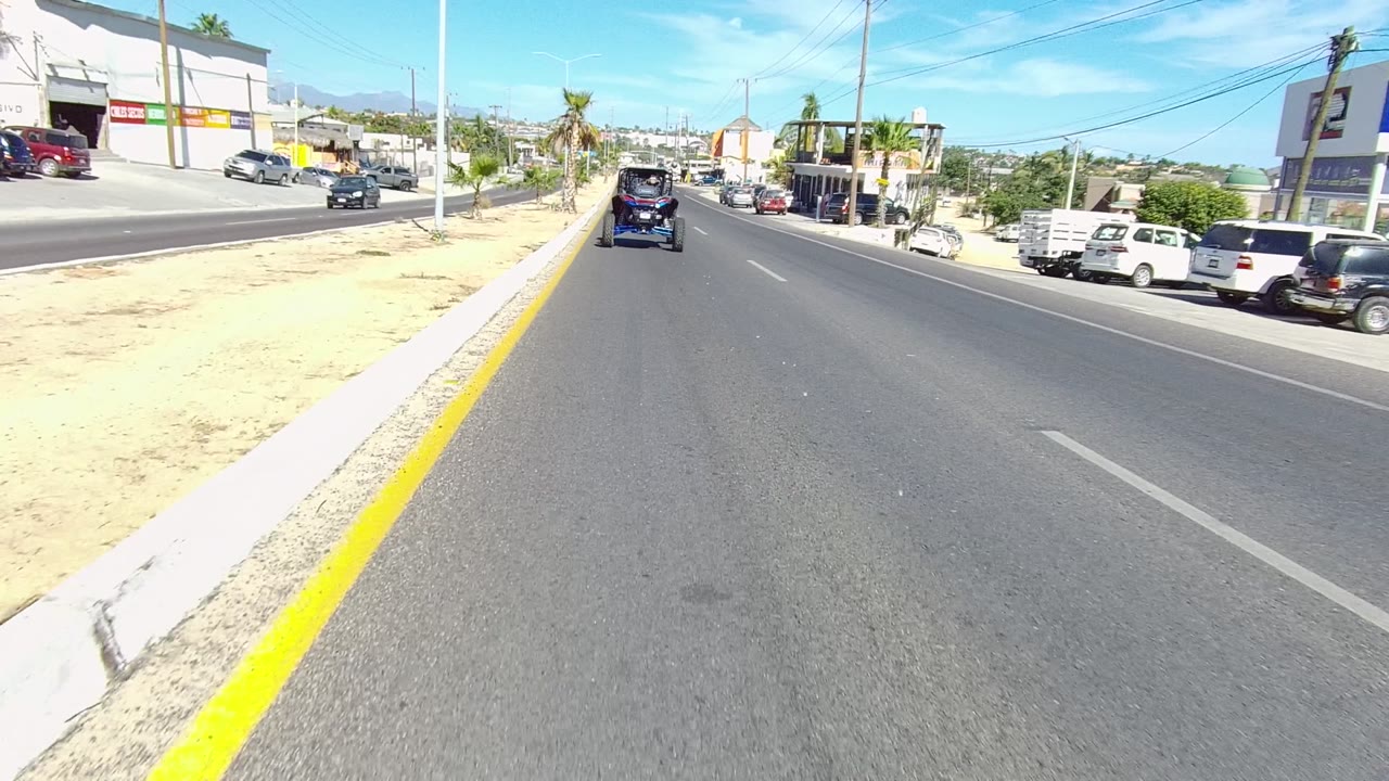 Golf cart centered in the lane with commercial buildings and parked vehicles along the road.