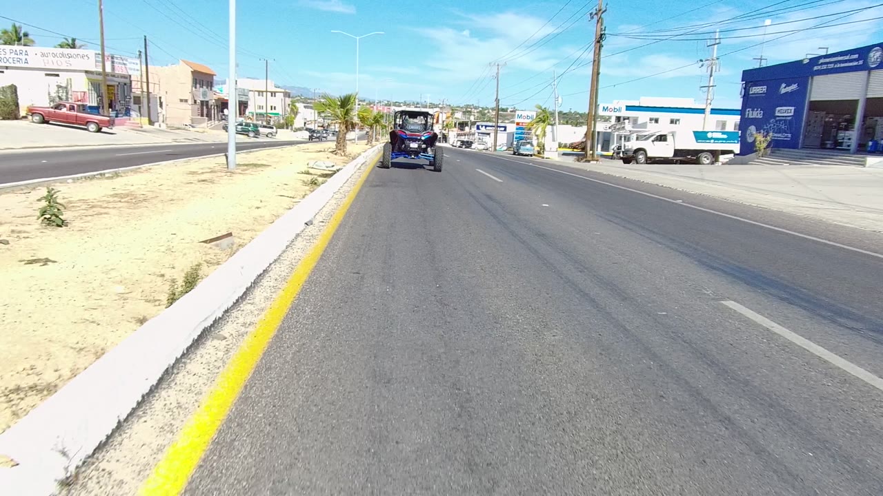 Opening frame of a Los Cabos road with a yellow curb line on the left, sandy verge, palm trees, distant cars, utility wires, commercial buildings, and no visible people.