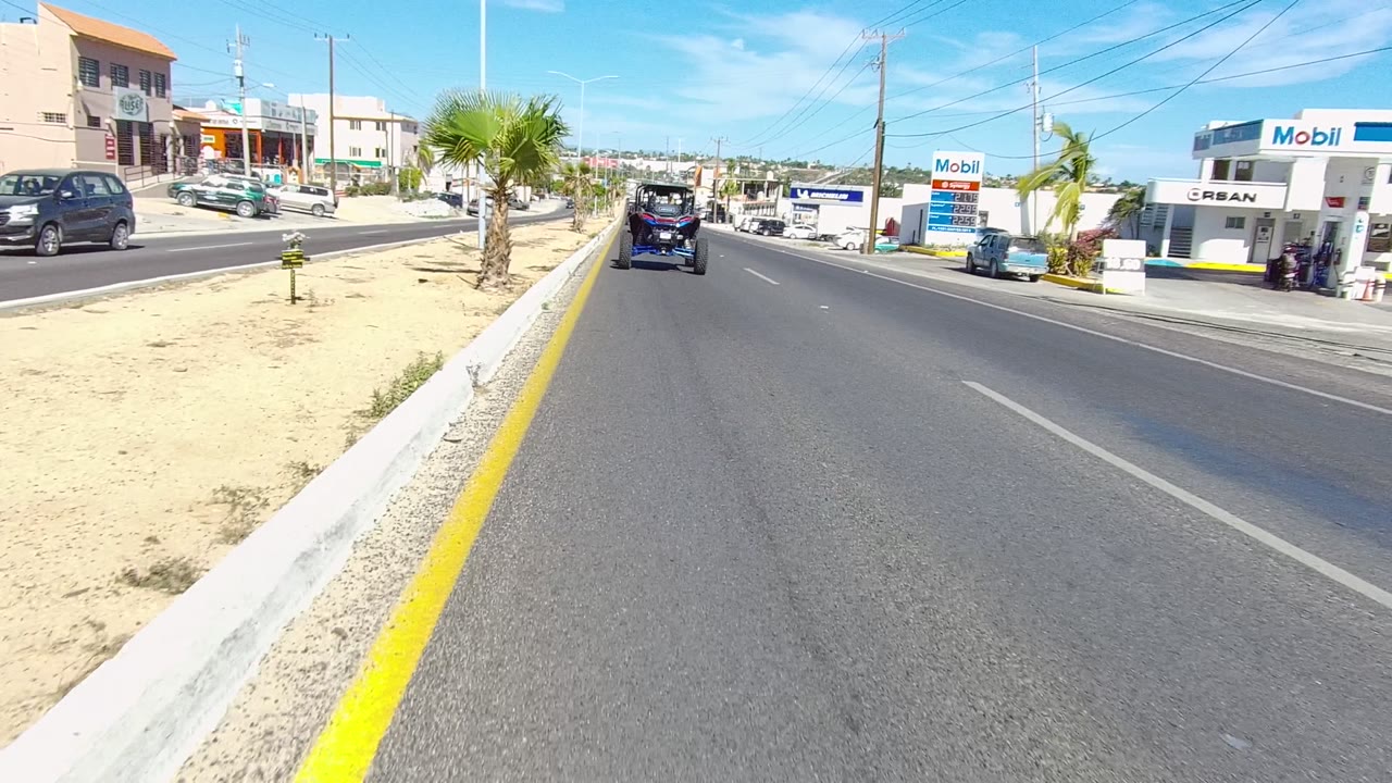 Mid-clip Los Cabos road frame with Mobil gas station signage on the right, parked vehicles near the curb, a palm in the median, overhead wires, and no visible people.