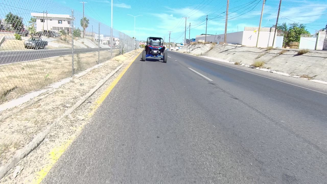 Road-level view in Los Cabos, Baja California Sur, with a yellow center line, chain-link fence, low buildings, utility poles, dry roadside ground, a vehicle ahead, and no visible people.