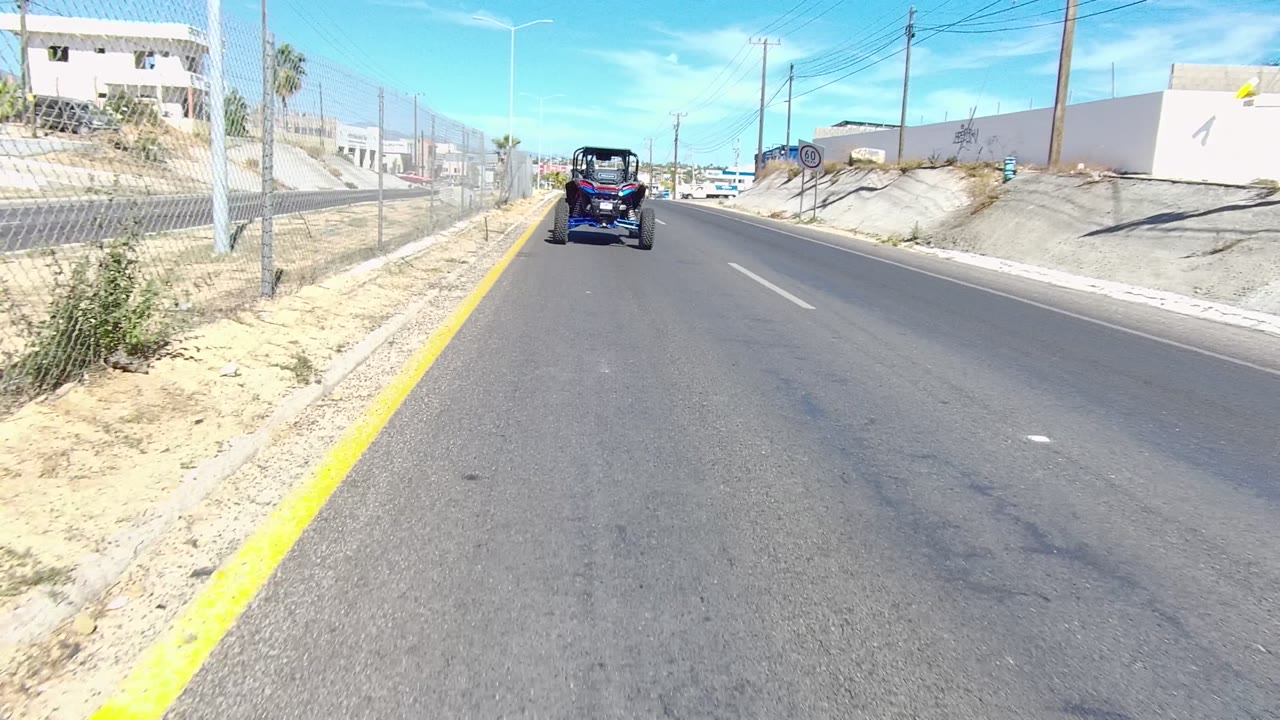 Straight paved road beside dry shoulder, fence, buildings, and a vehicle ahead.