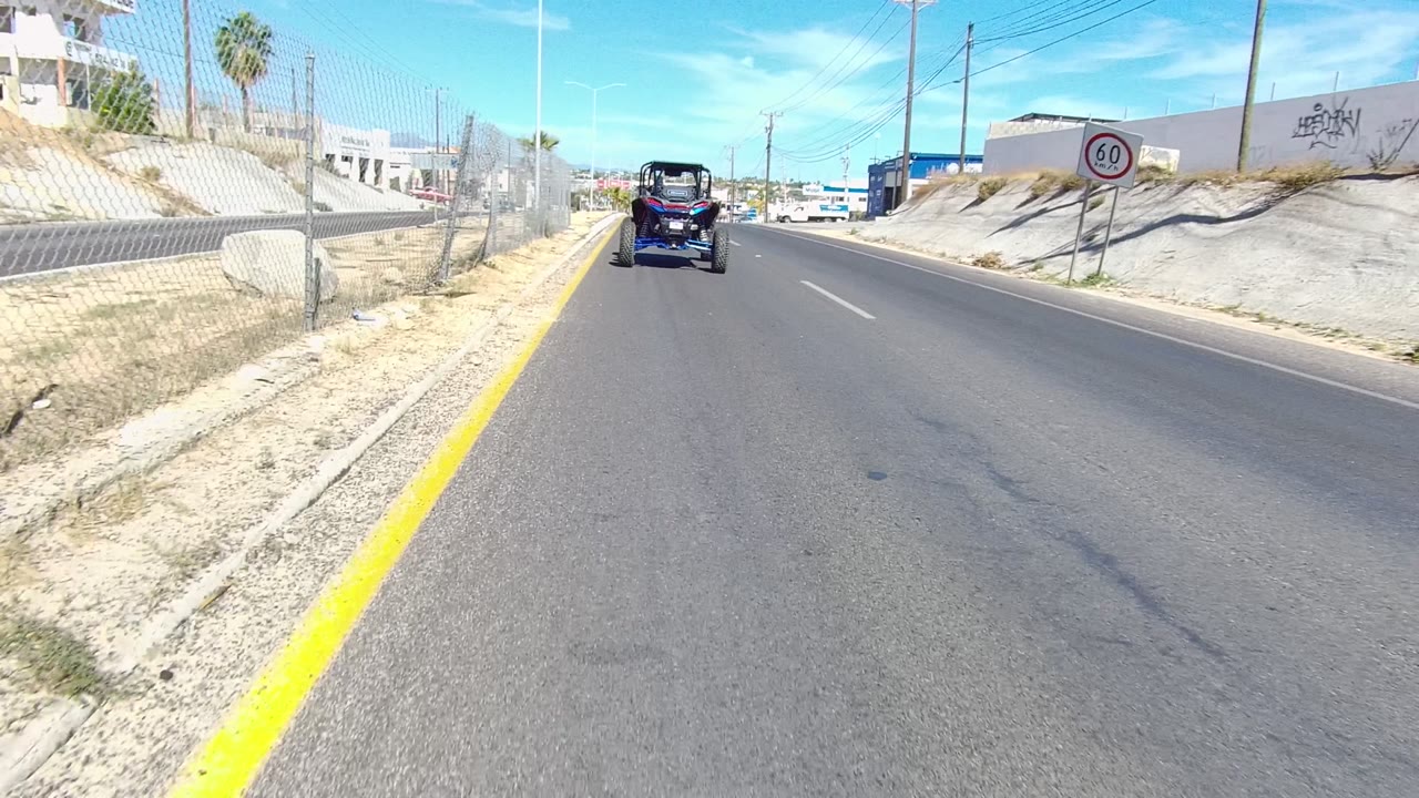 At 0:02, a circular speed sign appears on the right side of the Los Cabos road, with a dry sloped embankment, utility lines, low buildings, a vehicle ahead, and no visible people.