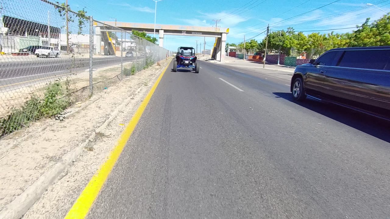 Road-level view in Los Cabos, Baja California Sur showing a yellow curb line, chain-link fence, small vehicle ahead, distant overpass, blue sky, and no visible people.