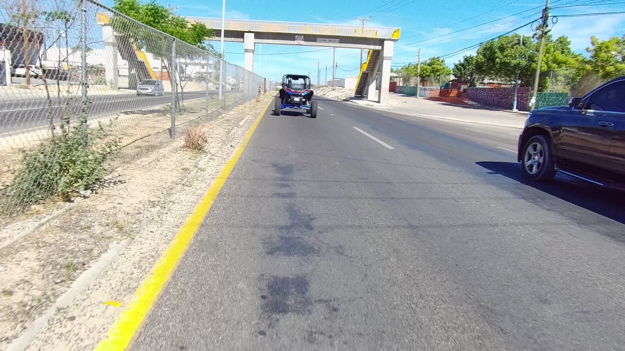 Paved lane beside a chain-link fence with a small vehicle ahead and a dark SUV along the right edge.