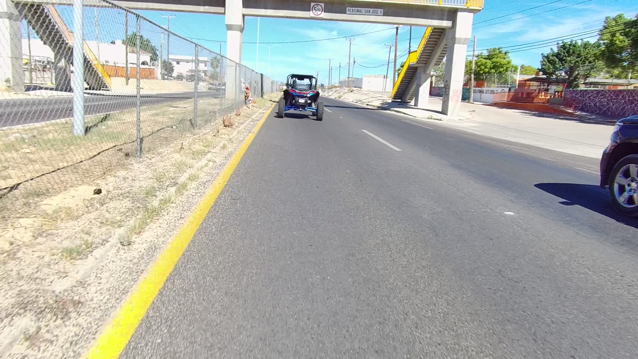 Bright Los Cabos street view approaching the overpass, with dry roadside ground, chain-link fence, yellow curb line, vehicle ahead, partial SUV at right, and no visible people.
