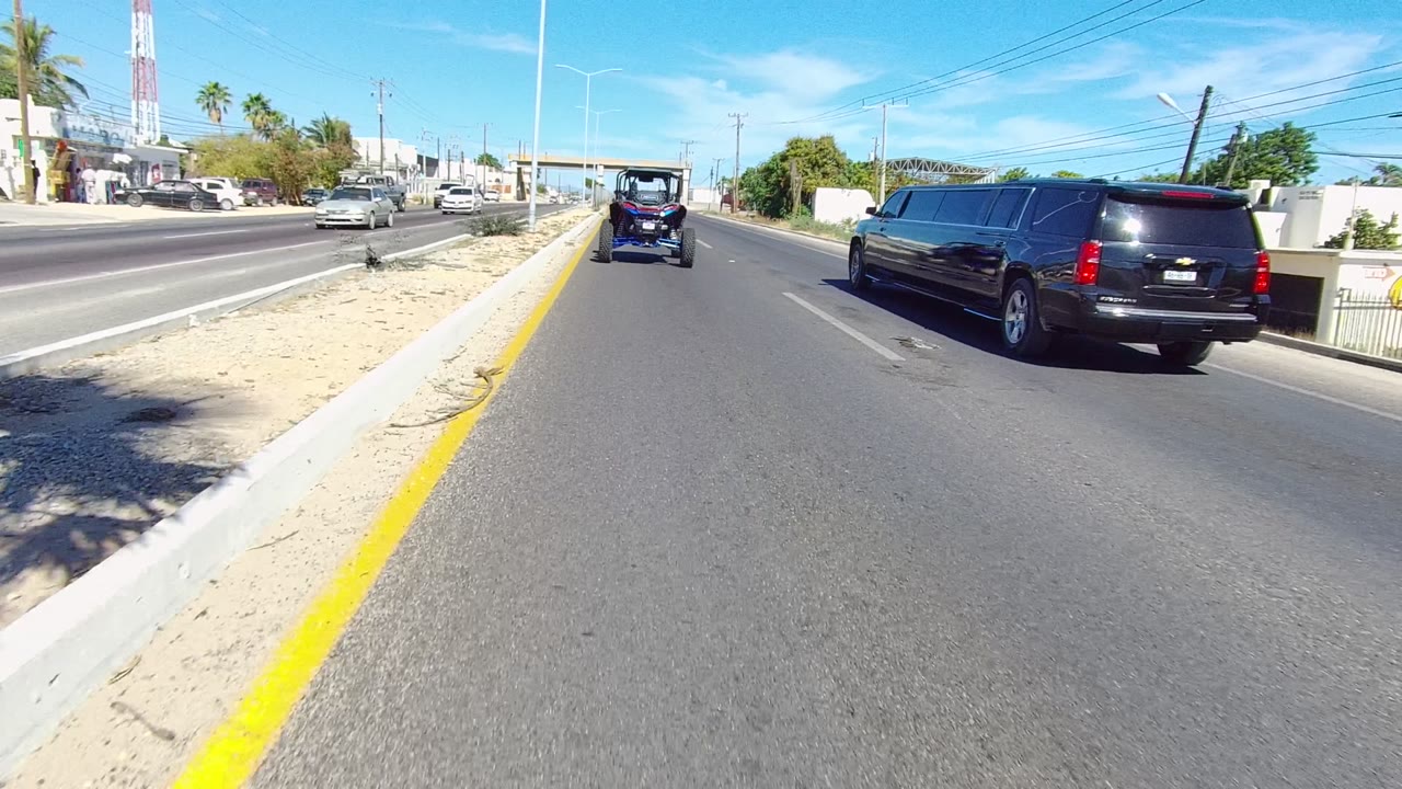 At 0:00 in Los Cabos, a black limousine follows a small golf cart along a bright multi-lane road with a yellow line, sandy median, palm trees, and no visible people.