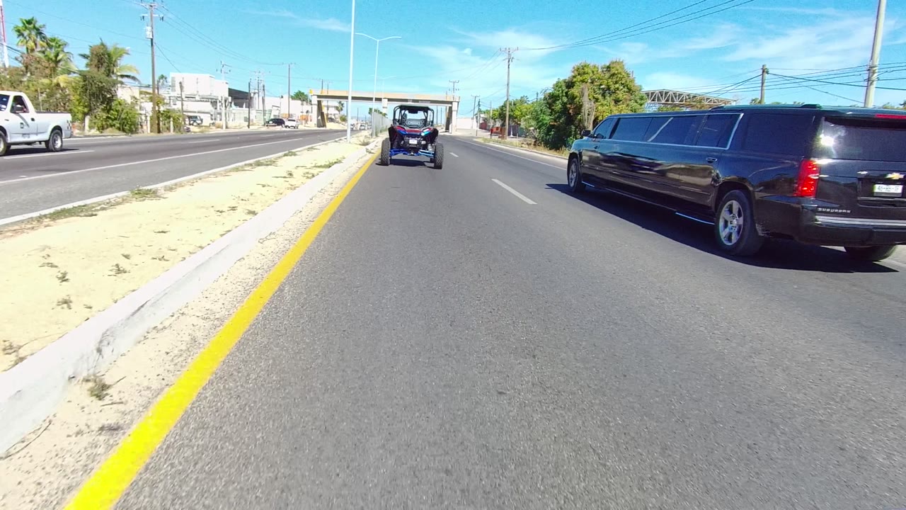 At 0:02 in Los Cabos, the limousine occupies the right side of the frame while the golf cart remains centered ahead, with oncoming vehicles, a sandy divider, palms, and no visible people.