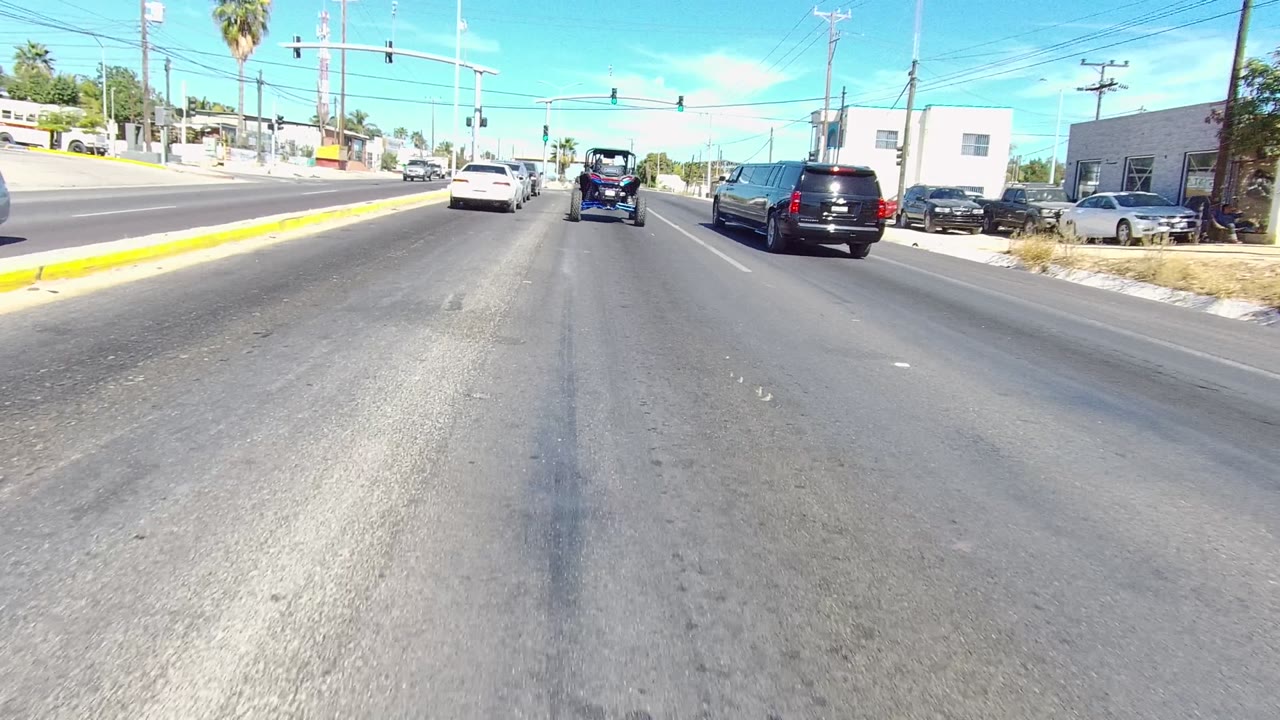 Street-level view of traffic on a multi-lane road in Los Cabos, Baja California Sur, with a small blue vehicle ahead, cars in adjacent lanes, traffic lights, low buildings, palms, and no visible people.