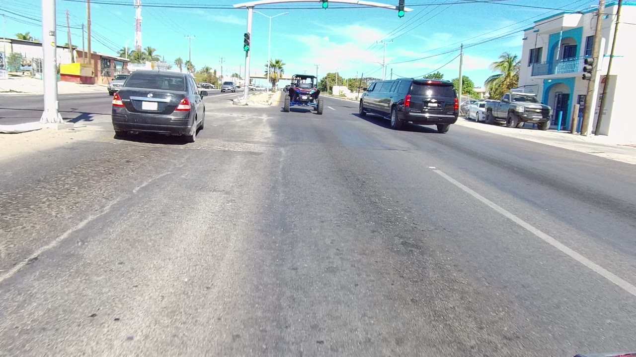 Centered roadway view in Los Cabos with a black SUV ahead, a small blue vehicle farther forward, signal lights overhead, curbside buildings, palms, utility lines, and no visible people.