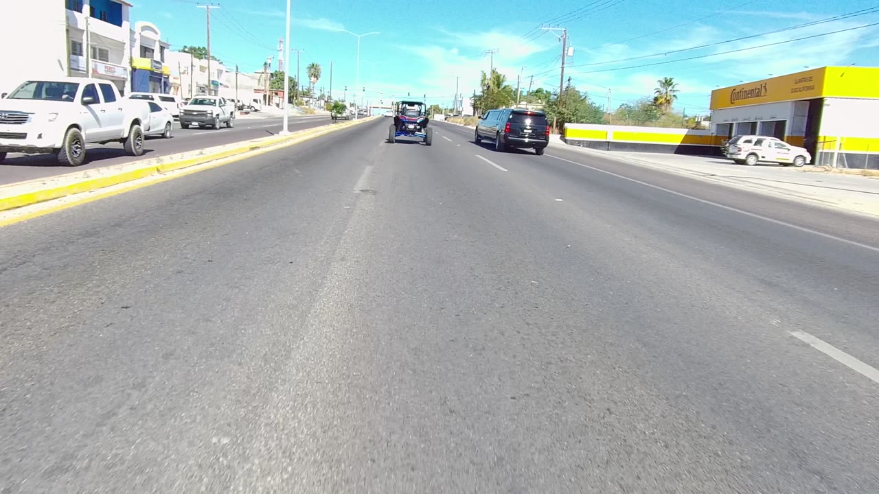 Opening roadway view in Los Cabos, Baja California Sur with cars ahead in multiple lanes, a yellow curb median, low buildings, palm trees, and no visible people.