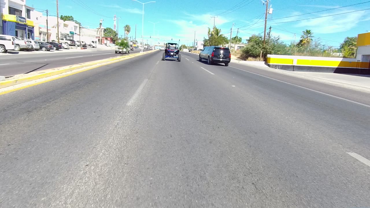 Urban road with vehicles ahead, a yellow building at right, sidewalks, palms, and clear sky.