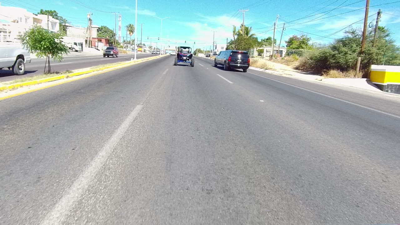 Two-second frame from the Los Cabos road clip with cars ahead, a white van at the right roadside, small buildings, trees, wires, and no visible people.