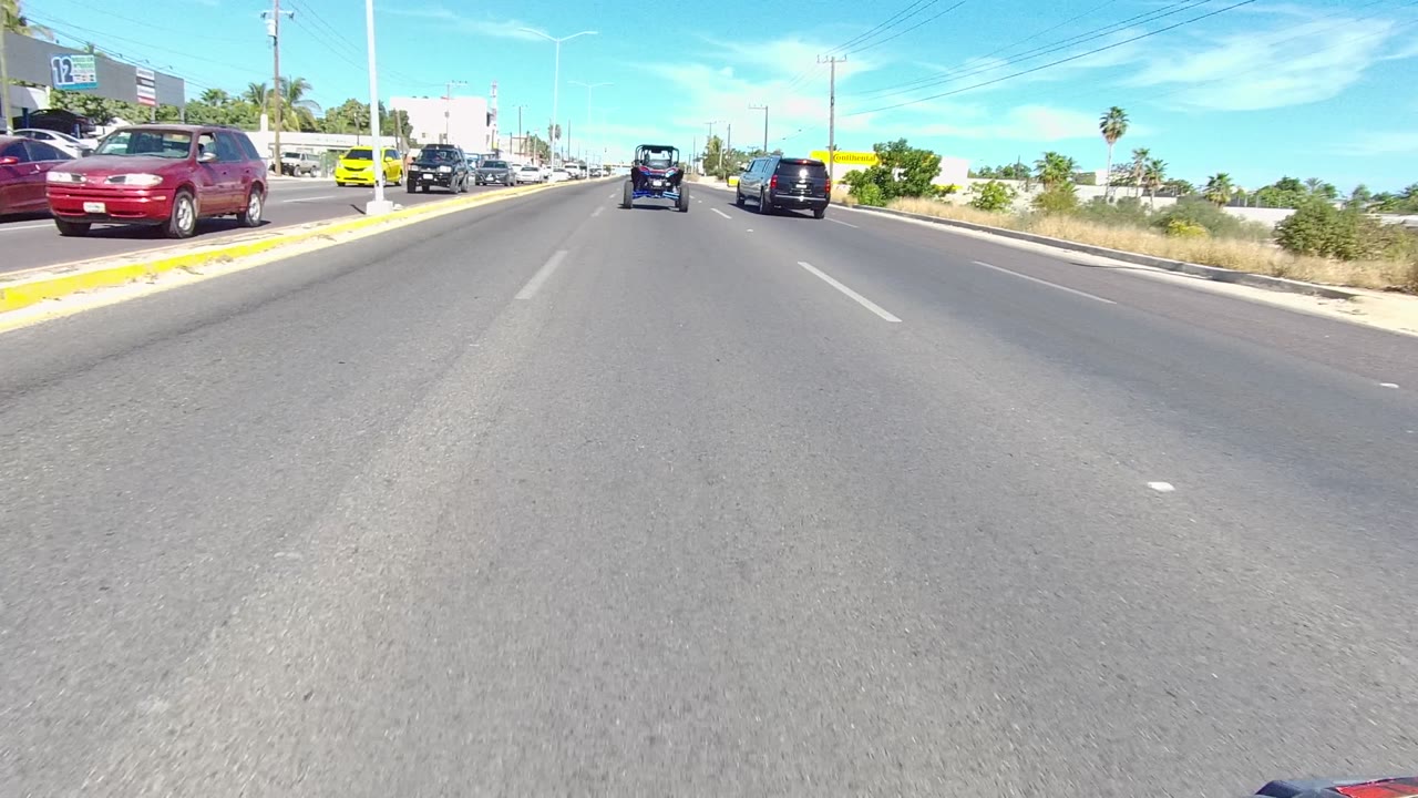 Forward road view in Los Cabos, Baja California Sur with cars ahead, passing traffic at left, palm trees, low buildings, and no visible people.