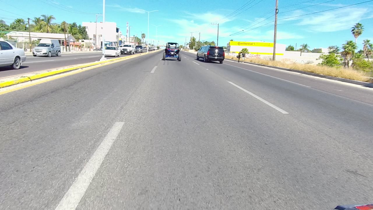A daytime Los Cabos traffic view shows open pavement, cars ahead in the same direction, oncoming vehicles at left, a yellow building at right, palm trees, and no visible people.