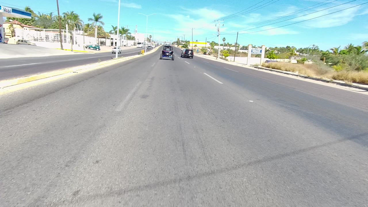 Start of the Los Cabos road clip showing cars ahead on a wide asphalt street, palm trees and low buildings on the left, utility lines overhead, and no visible people.