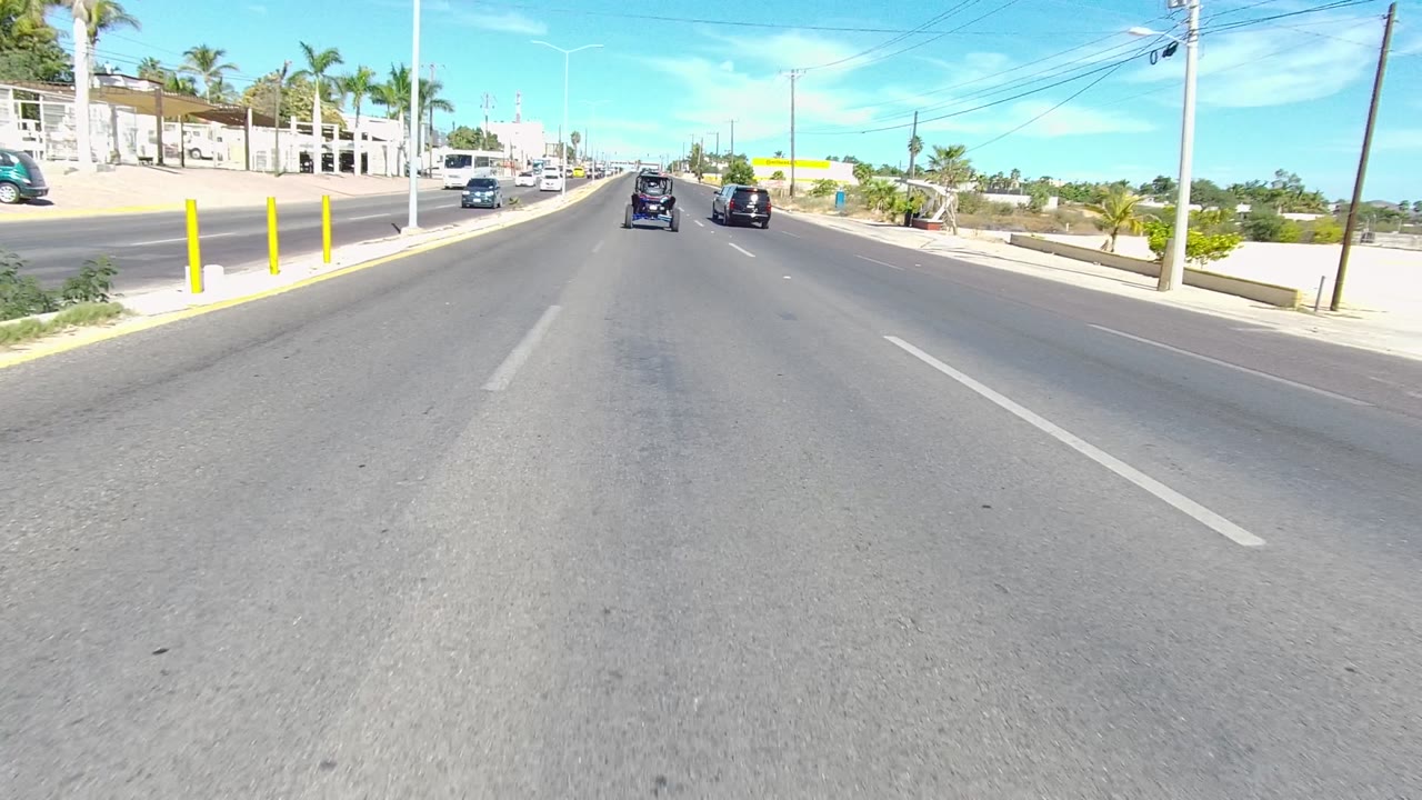 Two seconds into the Los Cabos driving view, two vehicles remain ahead in the lanes while palm trees, low commercial buildings, and utility poles line the bright roadside, with no visible people.